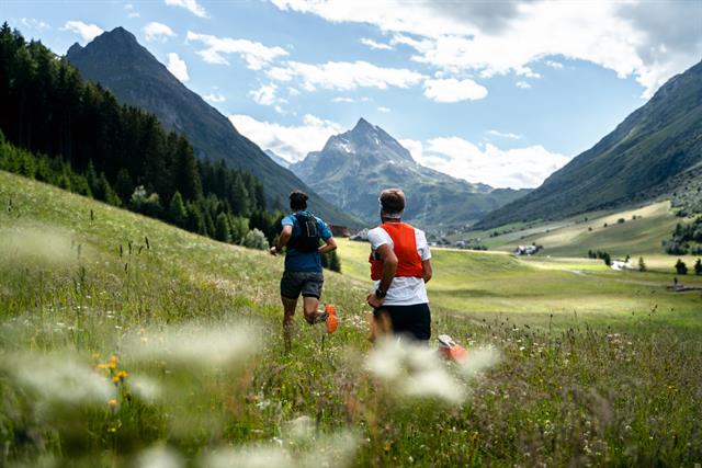Zwei Trailrunner sprinten über eine grüne Wiese, umgeben von majestätischen Berggipfeln